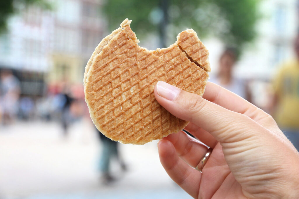 Hand mit angebissener Stroopwafel vor verschwommenem Amsterdamer Stadthintergrund