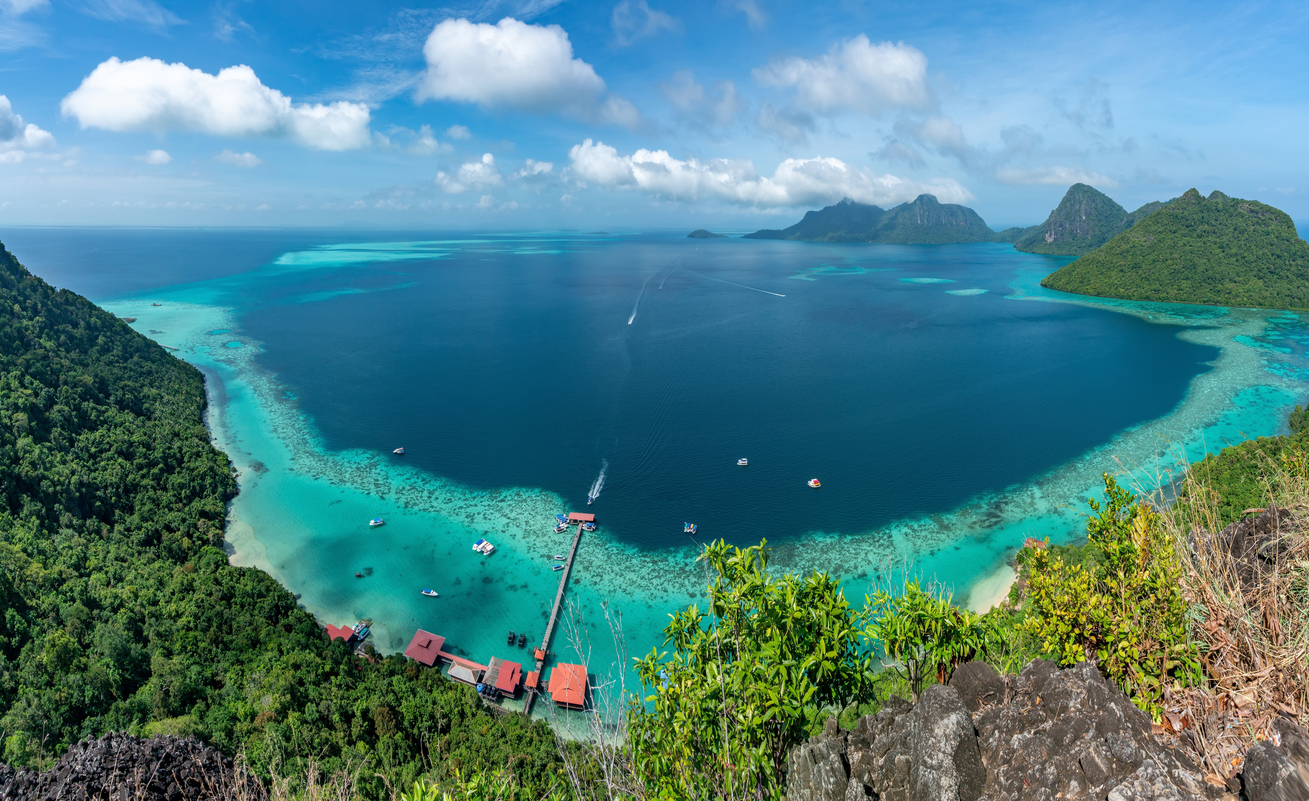 Panoramablick auf Lagune in Sabah mit Korallenriffen, Steg mit roten Dächern, Booten und bewaldeten Inseln