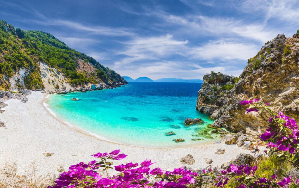 Türkisblaue Bucht auf Lefkada mit Sandstrand, Felsen, grüner Vegetation und pinker Bougainvillea.