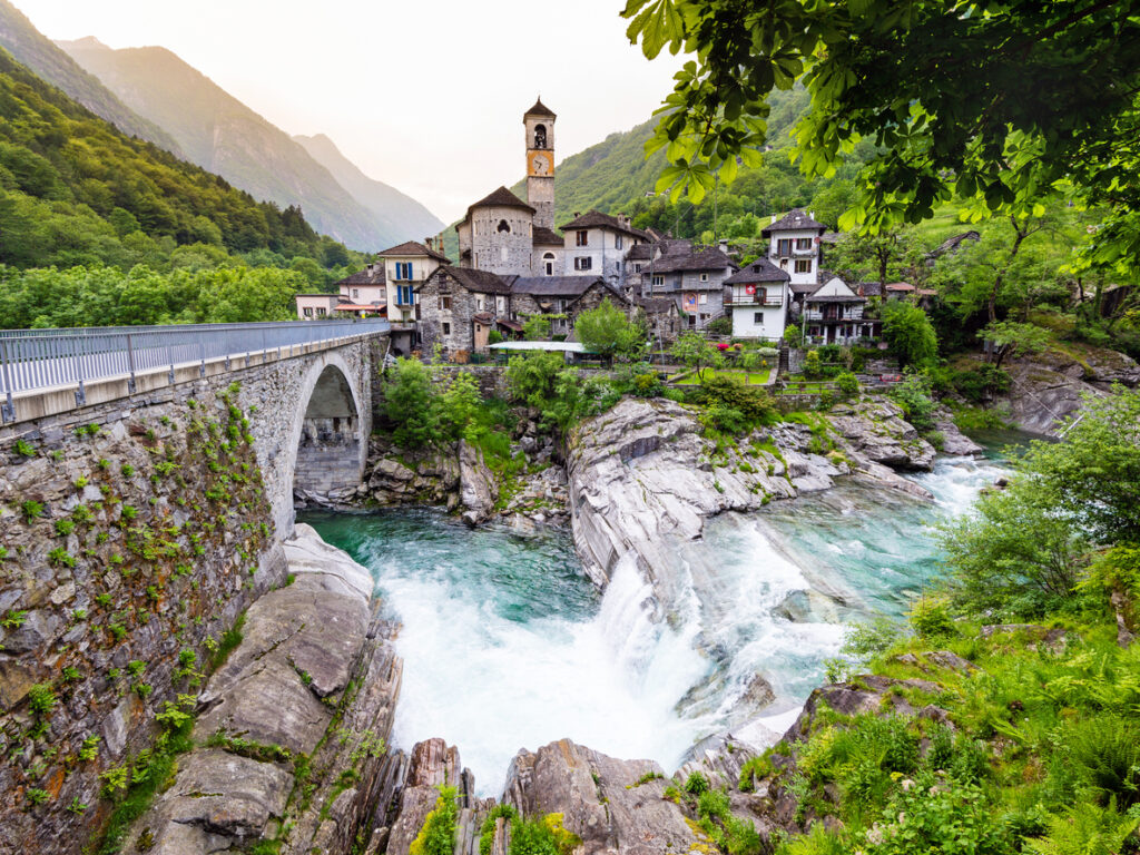 Val Verzasca mit Steinbrücke, türkisfarbenem Fluss und Wasserfall vor einem Bergdorf mit Kirchturm und bewaldeten Hängen