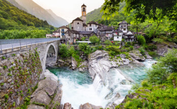 Val Verzasca mit Steinbrücke, türkisfarbenem Fluss und Wasserfall vor einem Bergdorf mit Kirchturm und bewaldeten Hängen