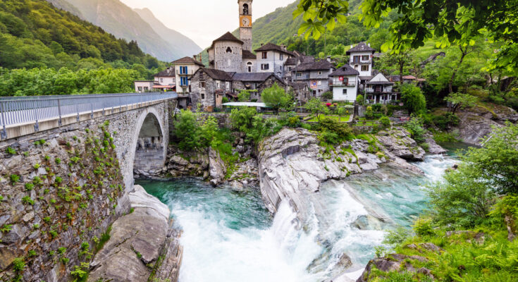 Val Verzasca mit Steinbrücke, türkisfarbenem Fluss und Wasserfall vor einem Bergdorf mit Kirchturm und bewaldeten Hängen