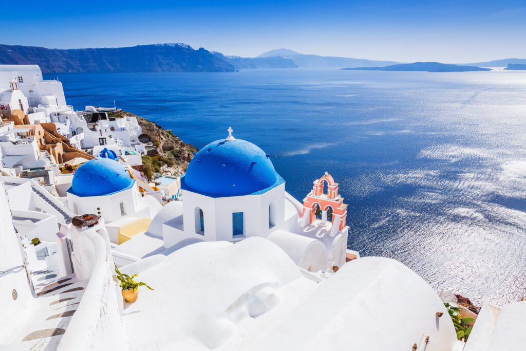 Blaue Kirchenkuppe und rosa Glockenturm in Oia, Santorini, mit Blick über die blaue Ägäis und Caldera.