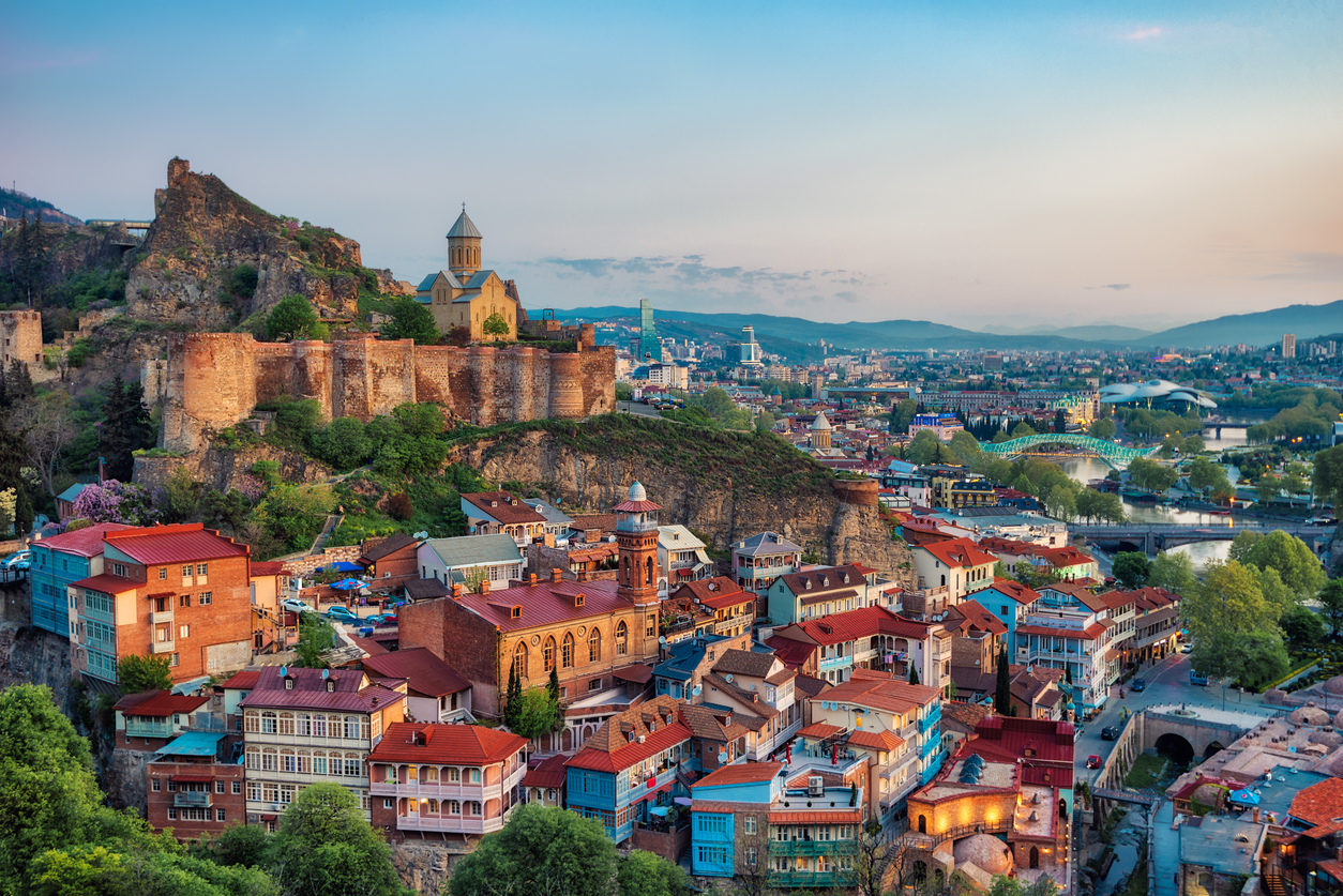 Panorama von Tiflis mit Narikala-Festung, Altstadthäusern und Friedensbrücke an der Kura bei Abenddämmerung.