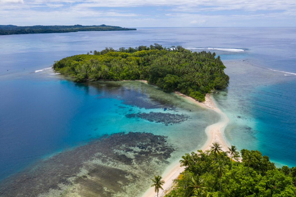 Luftaufnahme: bewaldete Inseln, helle Sandbank (Tombolo) und türkisfarbene Lagune mit Korallenriff.
