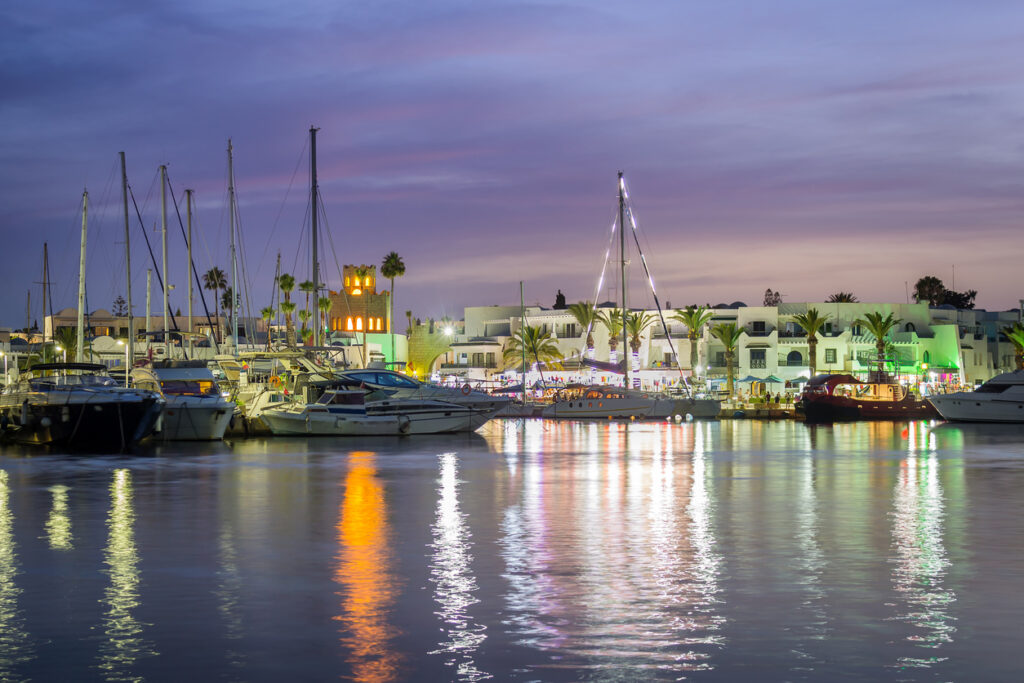 Abendstimmung im Yachthafen Port El Kantaoui mit beleuchteten Booten, Palmenpromenade und farbigen Reflexionen im ruhigen Mittelmeer.