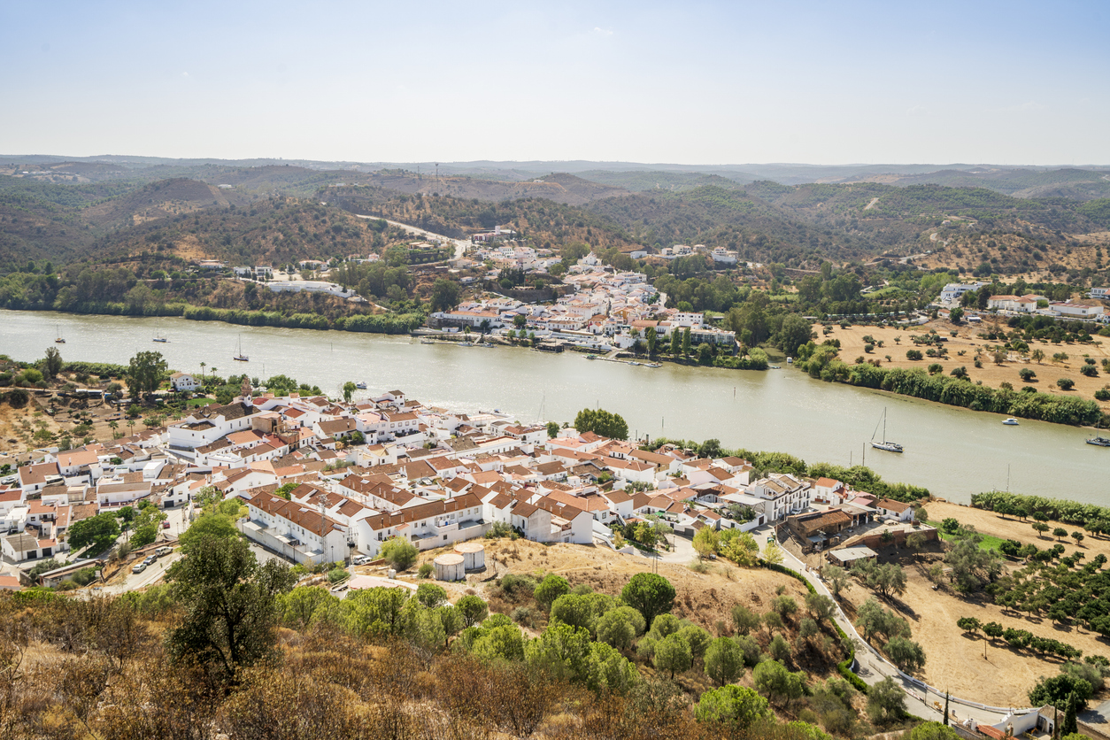 Blick auf Alcoutim am Fluss Guadiana mit weißen Häusern, Booten und hügeliger Landschaft.