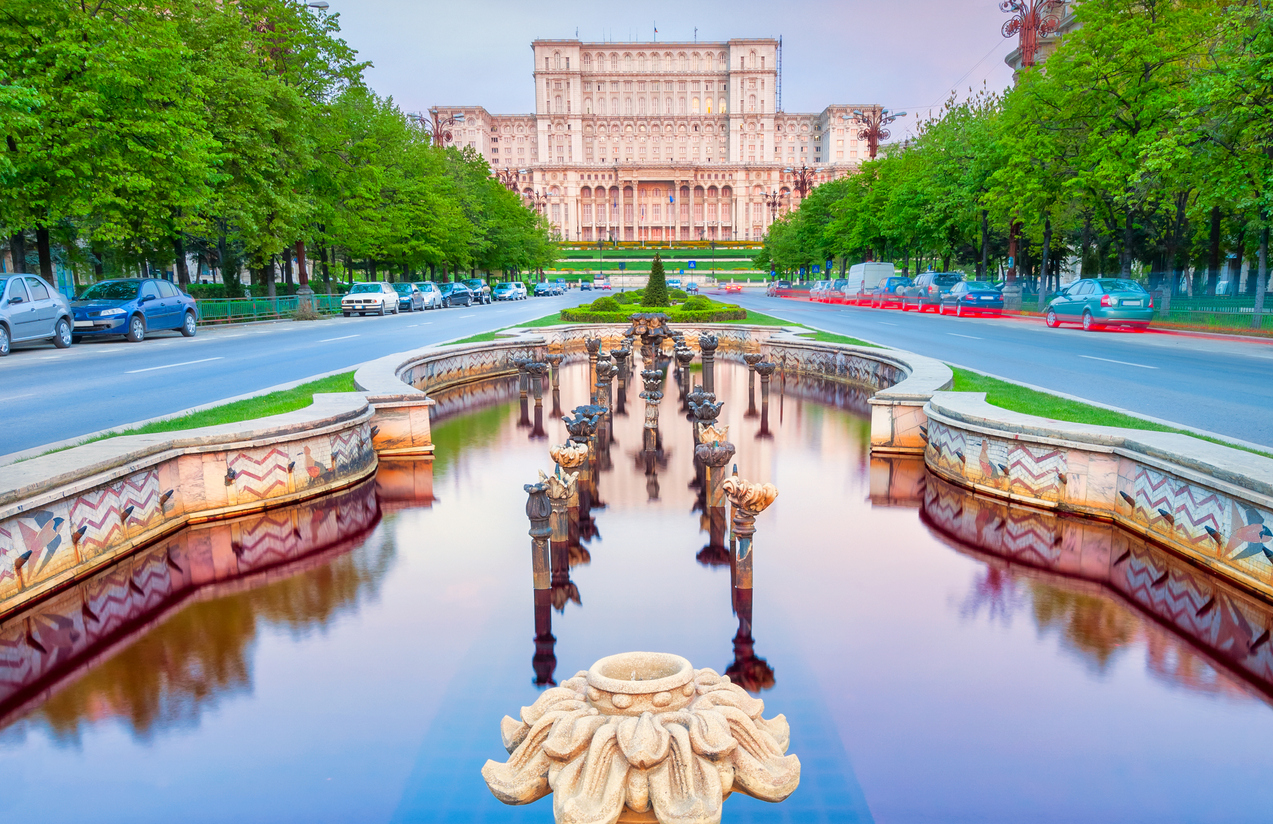 Blick auf das Parlamentsgebäude in Bukarest mit symmetrischem Brunnen im Vordergrund und von Bäumen gesäumter Straße.
