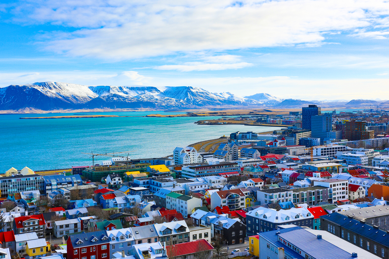Panorama von Reykjavík mit bunten Dächern, Bucht und schneebedeckten Bergen im Hintergrund.