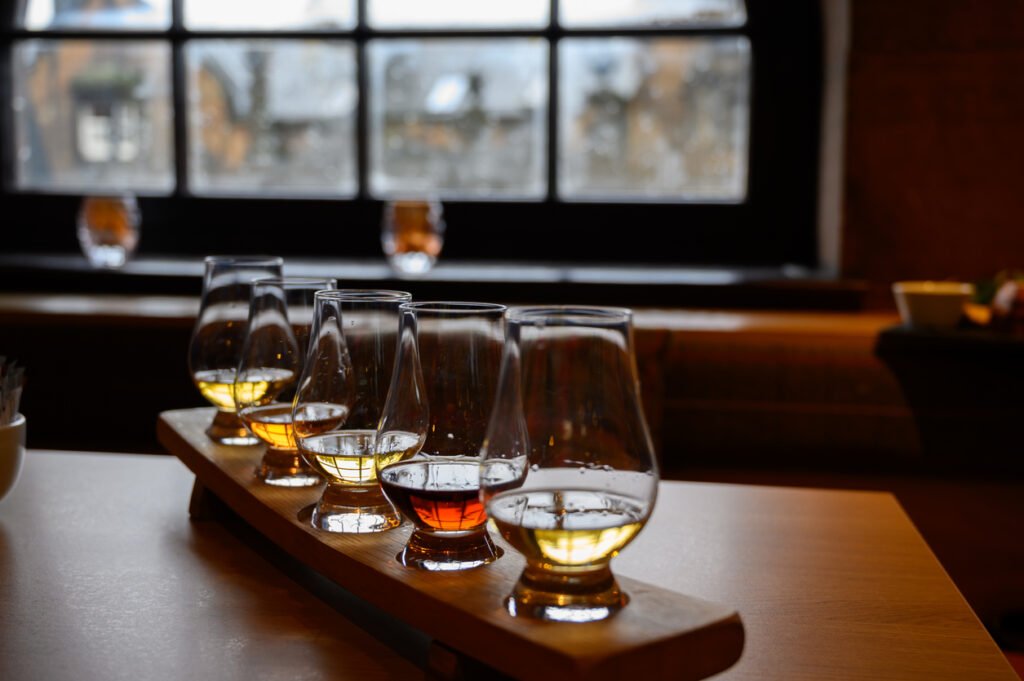 Fünf Glencairn-Gläser mit schottischem Whisky in unterschiedlichen Bernsteintönen auf einem hölzernen Probierbrett, Innenraum mit Fensterlicht, unscharfer Hintergrund