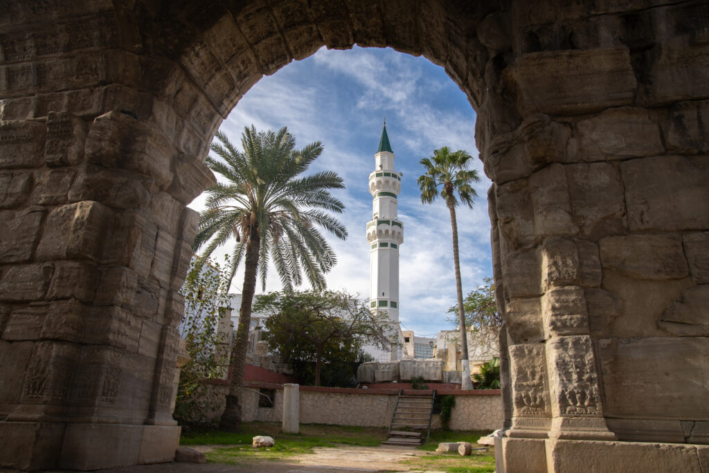 Durch antiken Steinbogen sichtbares weißes Minarett und Palmen in Tripolis, Libyen.