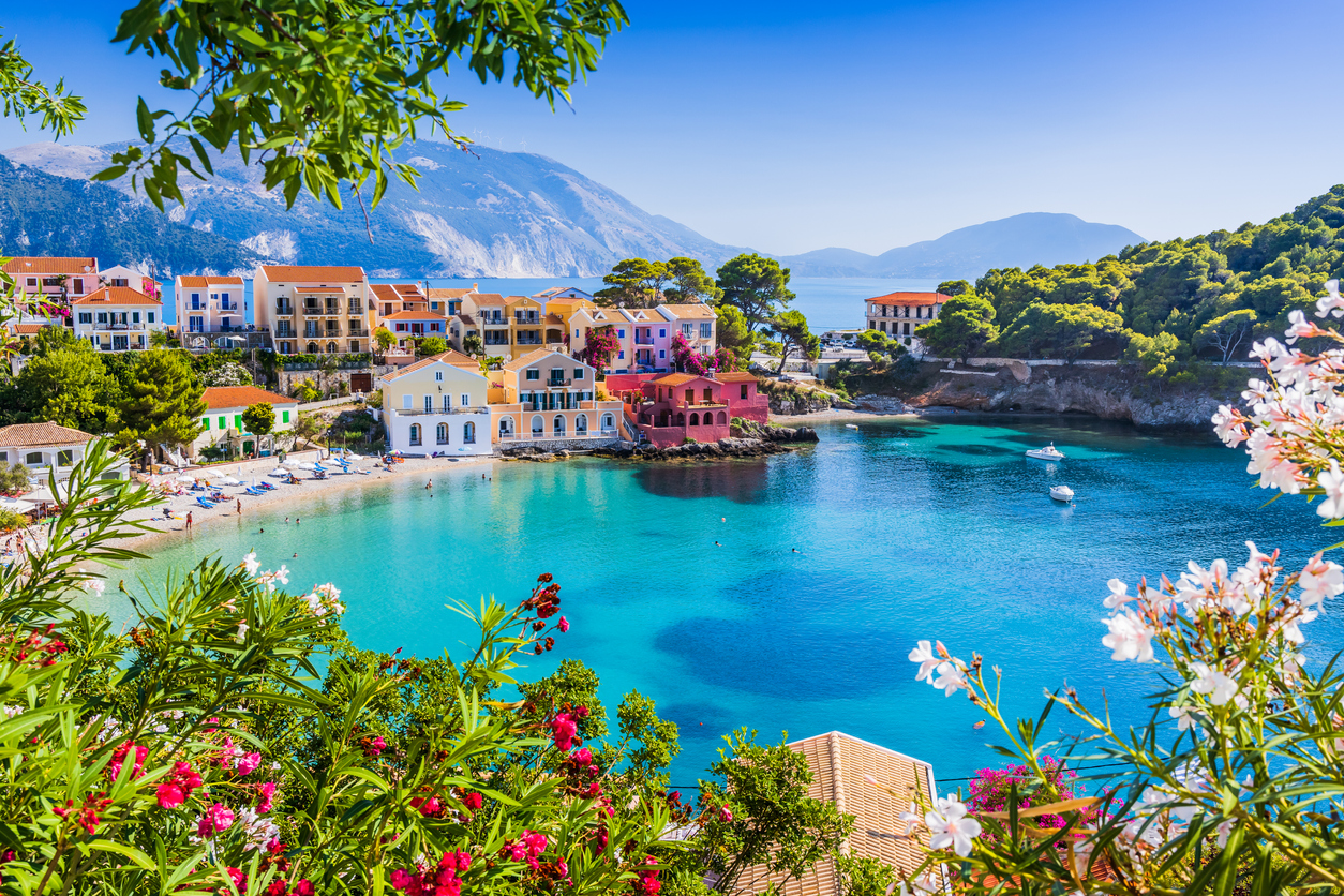 Bunte Küstenhäuser an einer türkisblauen Bucht auf Kefalonia, Blumen im Vordergrund, Boote und Berge.