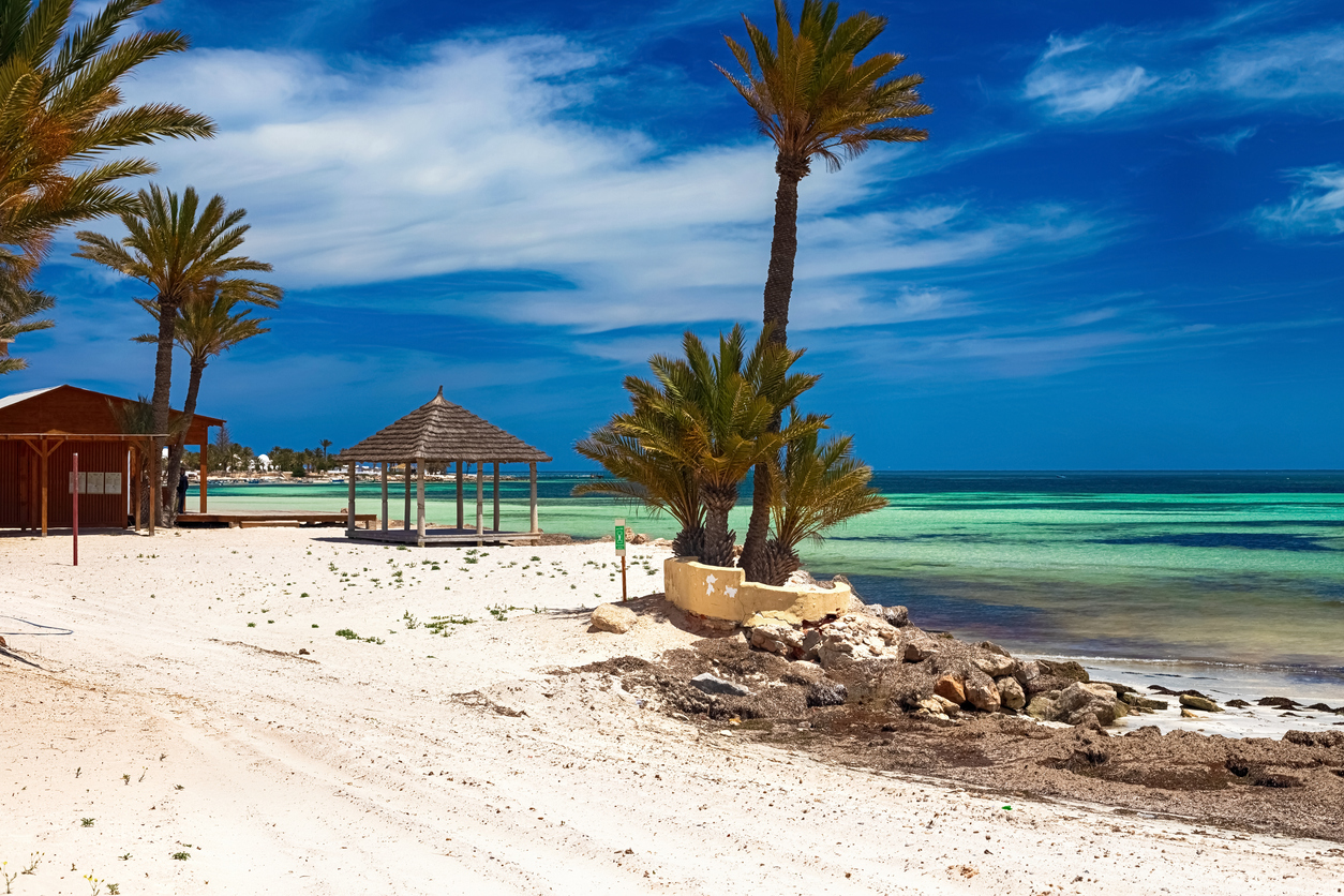 Heller Sandstrand auf Djerba mit Palmen, Holzpavillon und flachem, türkisblauem Mittelmeer unter tiefblauem Himmel.