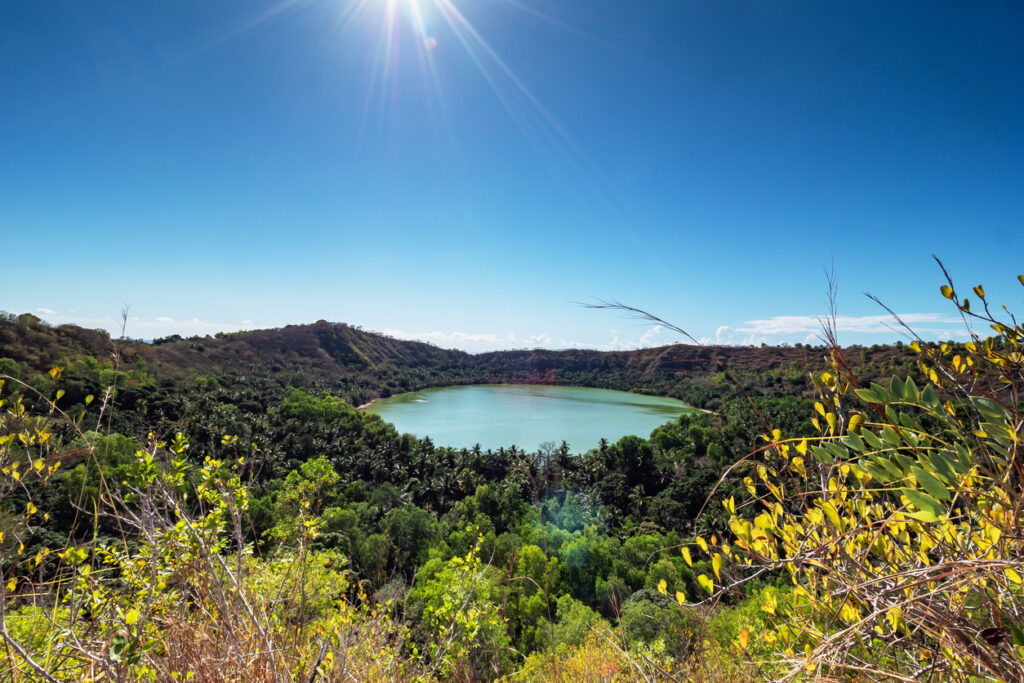 Grüner Kratersee auf den Komoren, von dichtem Regenwald und Hügeln umgeben, unter strahlender Sonne.