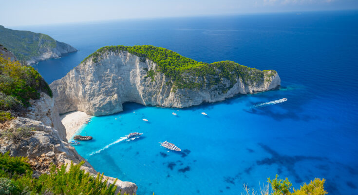 Türkisfarbene Bucht auf Zakynthos mit weißen Klippen, Sandstrand und Booten aus der Vogelperspektive.