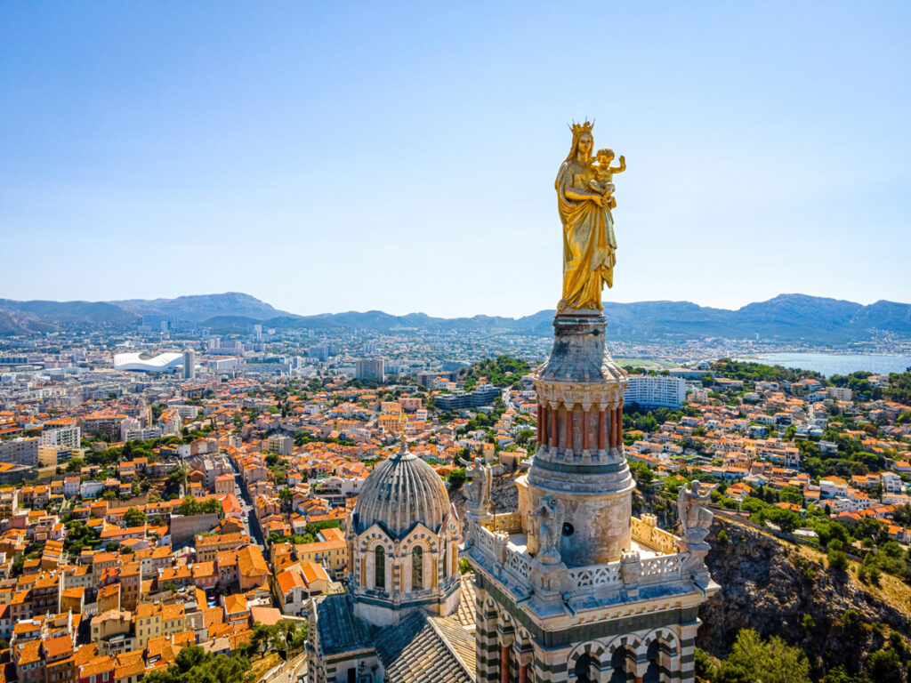 Goldene Marienstatue der Basilika Notre-Dame de la Garde ragt über den Dächern von Marseille, im Hintergrund Stadtpanorama, Berge und das glitzernde Mittelmeer bei klarem Himmel.
