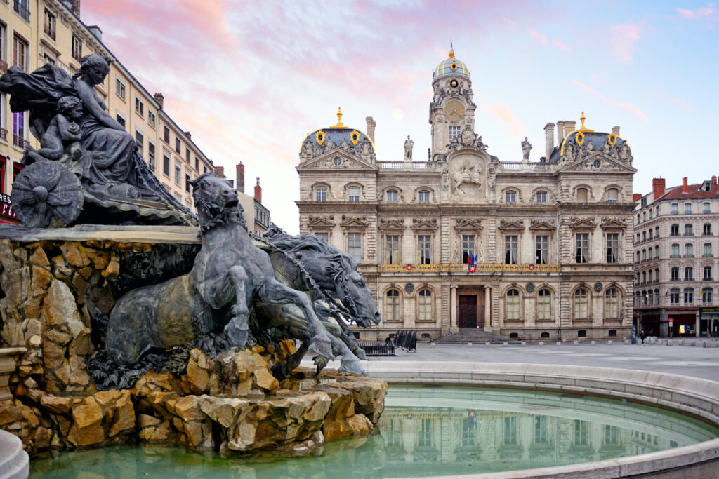 Weitwinkelaufnahme des Place des Terreaux in Lyon mit der bronzenen Fontaine Bartholdi im Vordergrund und dem prunkvollen Hôtel de Ville dahinter; ruhiger Platz, rosablauer Himmel.