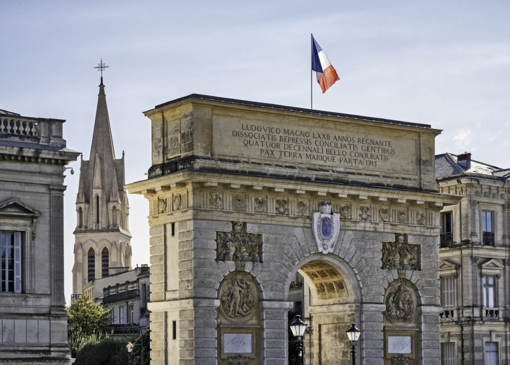 Steinener Triumphbogen Porte du Peyrou in Montpellier mit wehender französischer Flagge und lateinischer Inschrift; im Hintergrund ein neogotischer Kirchturm.