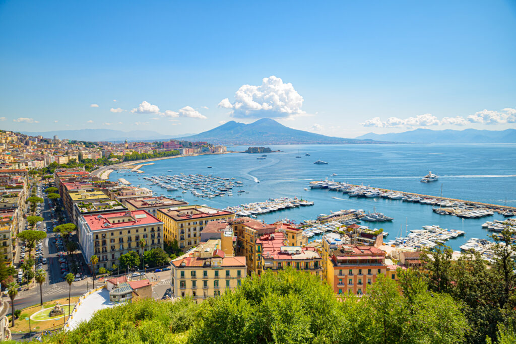 Neapel, Panoramablick über den Golf von Neapel mit Vesuv im Hintergrund, Uferpromenade mit dicht bebauten Vierteln, mehrere Marinas mit Yachten, Castel dell’Ovo auf Felseninsel, klarer Himmel