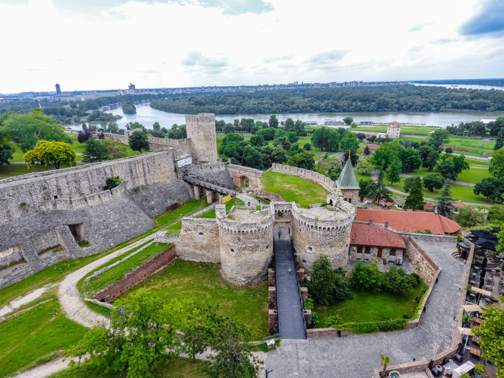 Belgrad, Luftaufnahme der Festung Kalemegdan mit runden Wehrtürmen, Toranlage und Brücke über den Graben, umgeben von Parkflächen; im Hintergrund Flusslandschaft von Save und Donau, bewölkter Himmel
