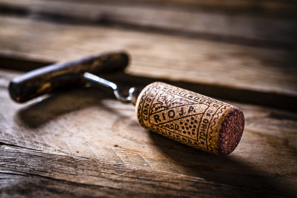 Makro eines Weinkorkens mit der Prägung „Rioja“, noch am Korkenzieher befestigt, liegend auf einem alten Holztisch; Fokus auf Struktur des Korks und weiche Lichtstimmung.