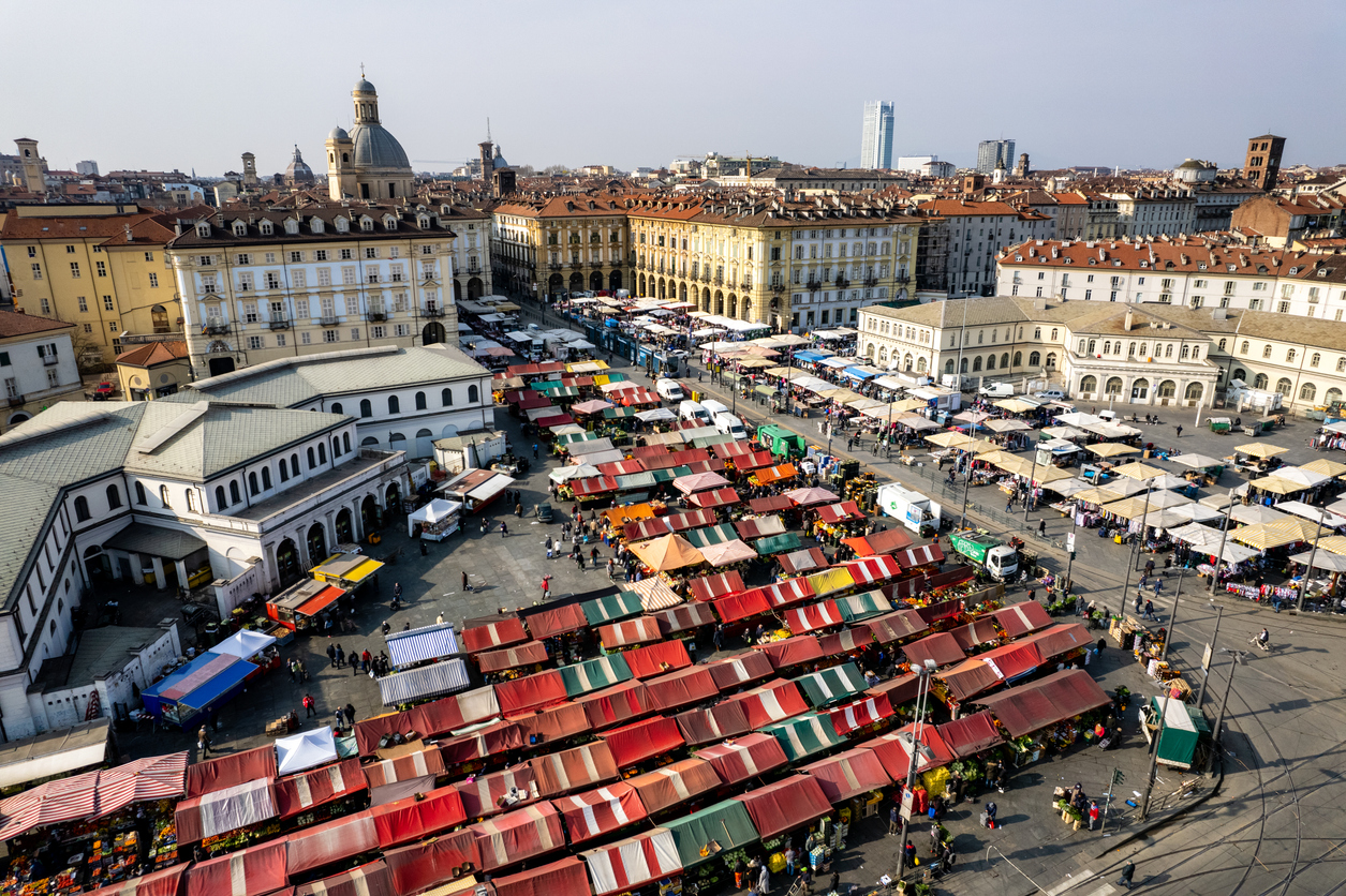 Luftaufnahme des Porta-Palazzo-Markts in Turin mit Reihen farbiger Marktdächer und vielen Ständen auf der Piazza della Repubblica, umgeben von historischen Gebäuden; im Hintergrund Kuppeln und ein moderner Turm der Stadt