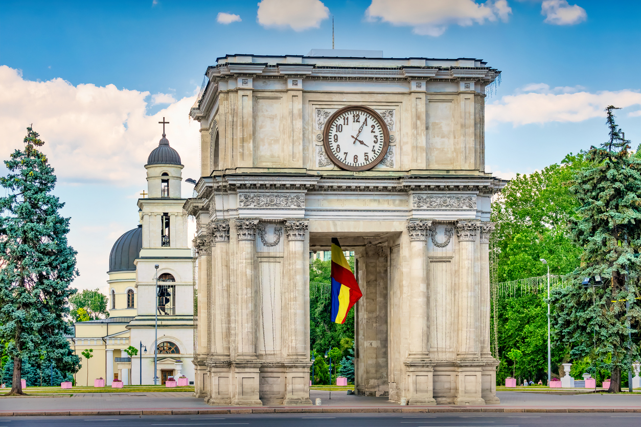 Frontalansicht des Triumphbogens (Arcul de Triumf) in Chișinău mit Uhr und wehender Flagge, dahinter der Kathedralglockenturm und grüne Bäume unter blauem Himmel.