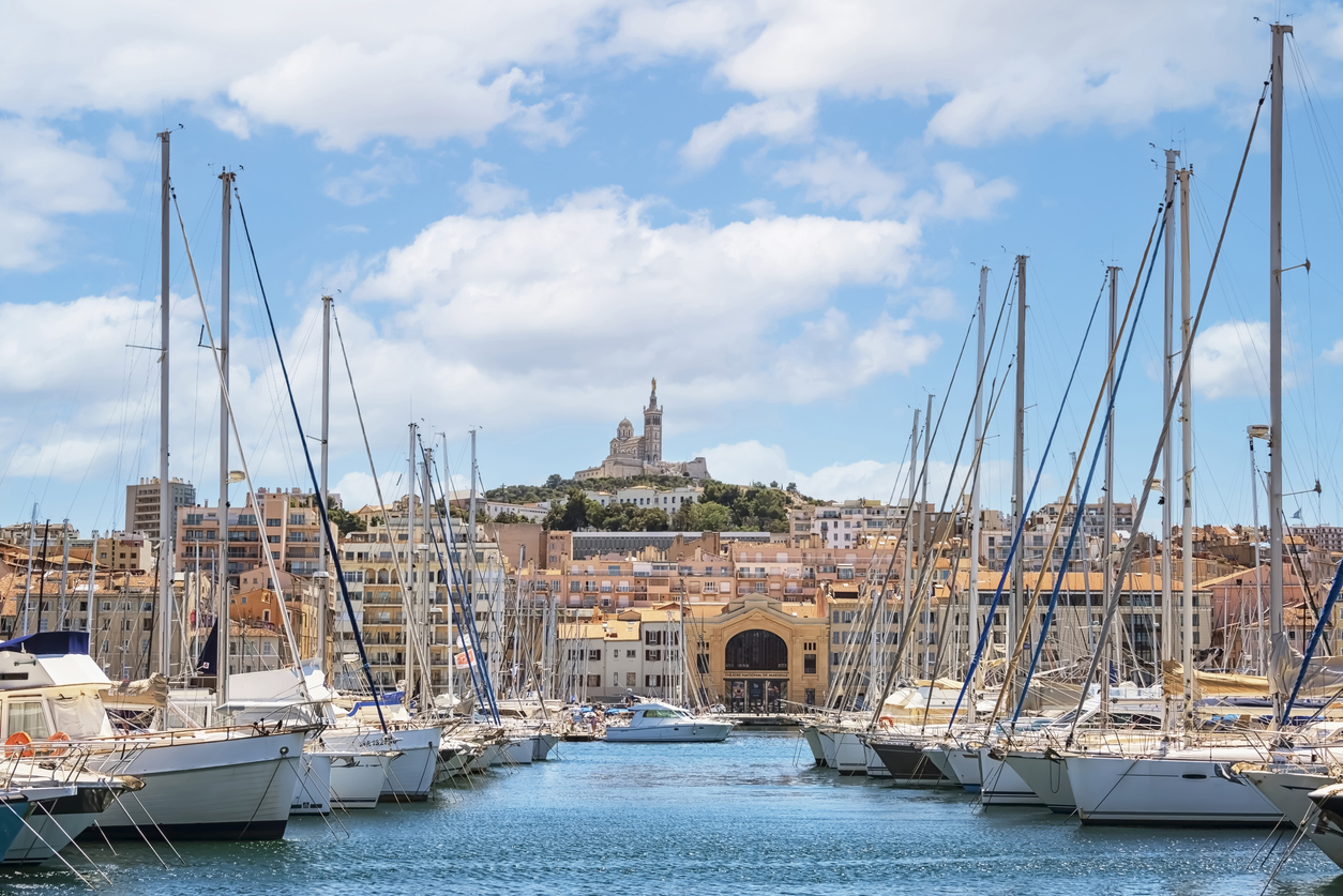 Blick durch Masten und Segelboote im Alten Hafen von Marseille auf pastellfarbene Häuser und die Basilika Notre-Dame de la Garde auf dem Hügel unter blauem Himmel.
