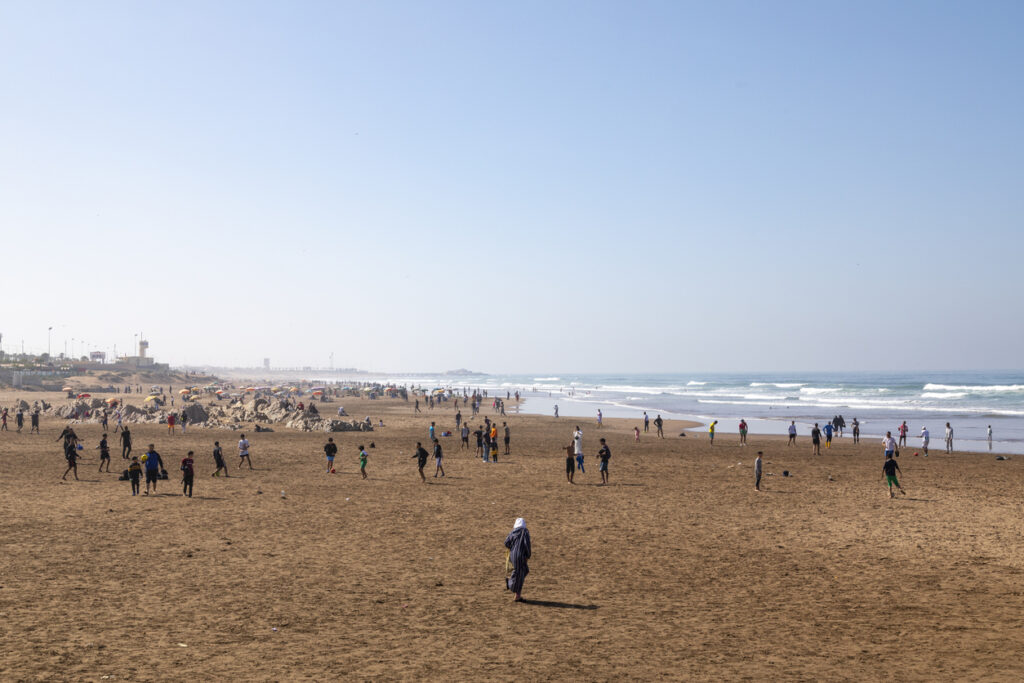 Belebter Sandstrand in Casablanca mit Fußballspielern, Sonnenschirmen und Atlantikwellen.
