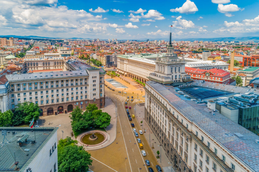 Sofia, Luftansicht des Largo mit turmgekröntem ehemaligen Parteigebäude rechts, neoklassizistische Amtsbauten, gelbe Pflasterstraßen, Glasdach über römischen Ruinen und Brunnen im Vordergrund, Sommerhimmel