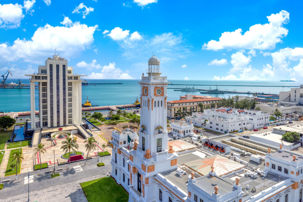 Weißer Uhrturm und Verwaltungsgebäude am Hafen von Veracruz; Meer und blauer Himmel mit Wolken.