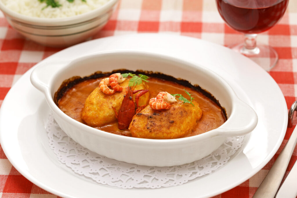 Zwei goldbraune Quenelles (Hechtklößchen) in cremiger Sauce Nantua, mit Flusskrebsen garniert, serviert in weißer Auflaufform auf rot-weiß karierter Tischdecke; daneben Rotweinglas und Reisschüssel im Hintergrund.