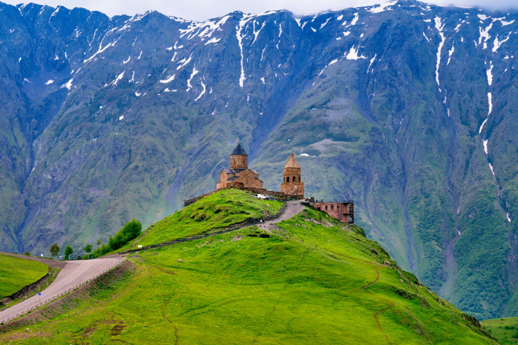 Gergeti-Dreifaltigkeitskirche auf grünem Hügel bei Kazbegi mit schneebedeckten Kaukasusbergen im Hintergrund.