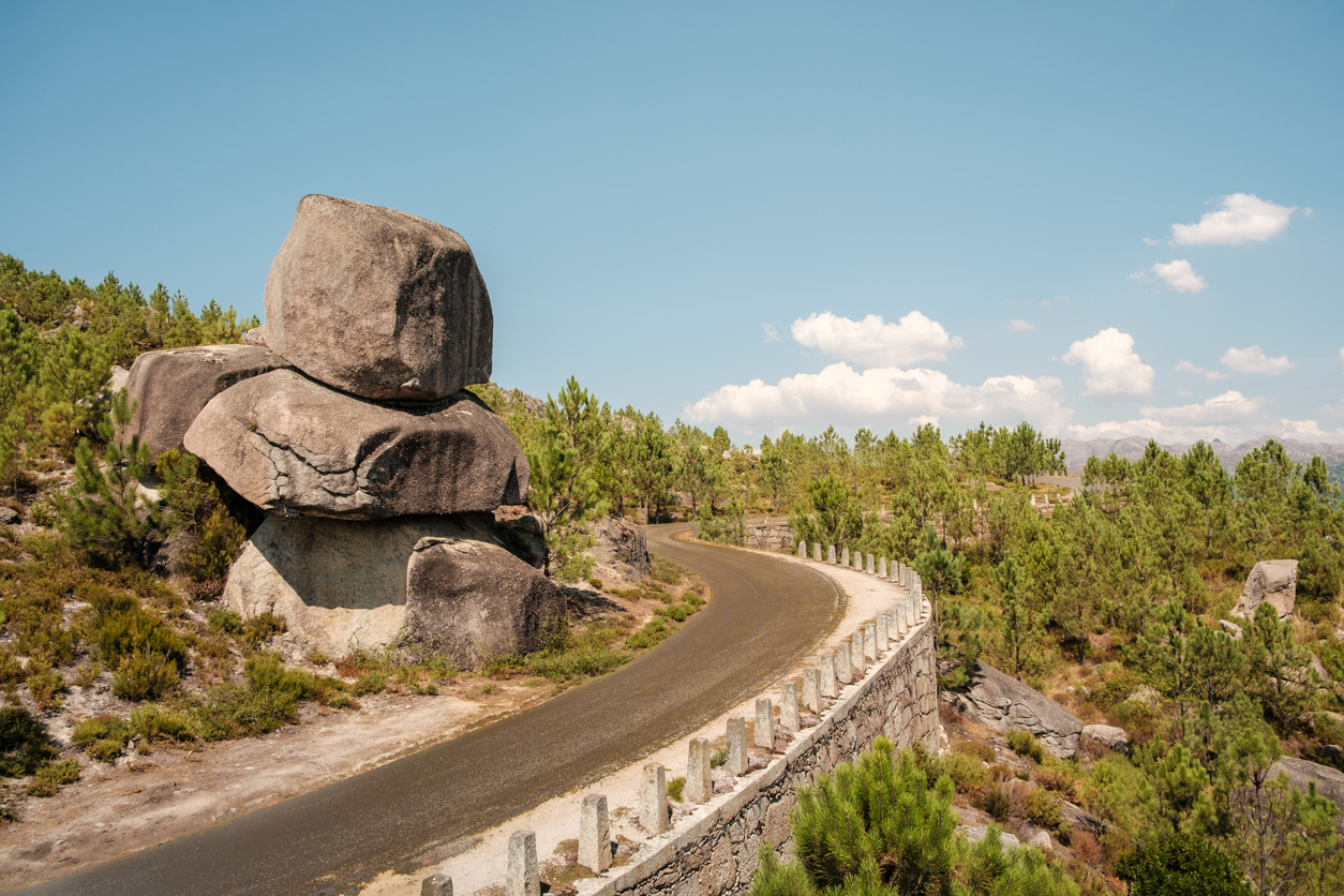 Kurvige Straße im Peneda-Gerês-Nationalpark neben gestapelten Granitfelsen und Kiefern, blauer Himmel.