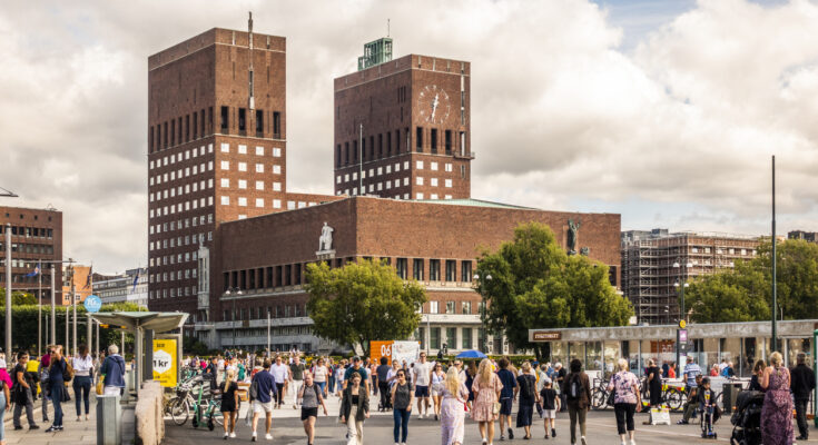 Menschen spazieren vor dem markanten Rathaus von Oslo mit seinen zwei Türmen.