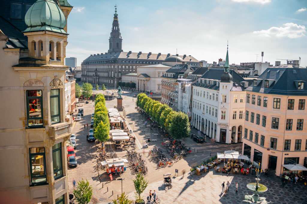 Blick auf die Strøget-Einkaufsstraße in Kopenhagen mit Cafés, Menschen und dem Christiansborg-Palast im Hintergrund.
