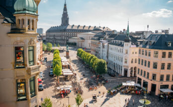 Blick auf die Strøget-Einkaufsstraße in Kopenhagen mit Cafés, Menschen und dem Christiansborg-Palast im Hintergrund.