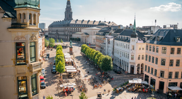 Blick auf die Strøget-Einkaufsstraße in Kopenhagen mit Cafés, Menschen und dem Christiansborg-Palast im Hintergrund.