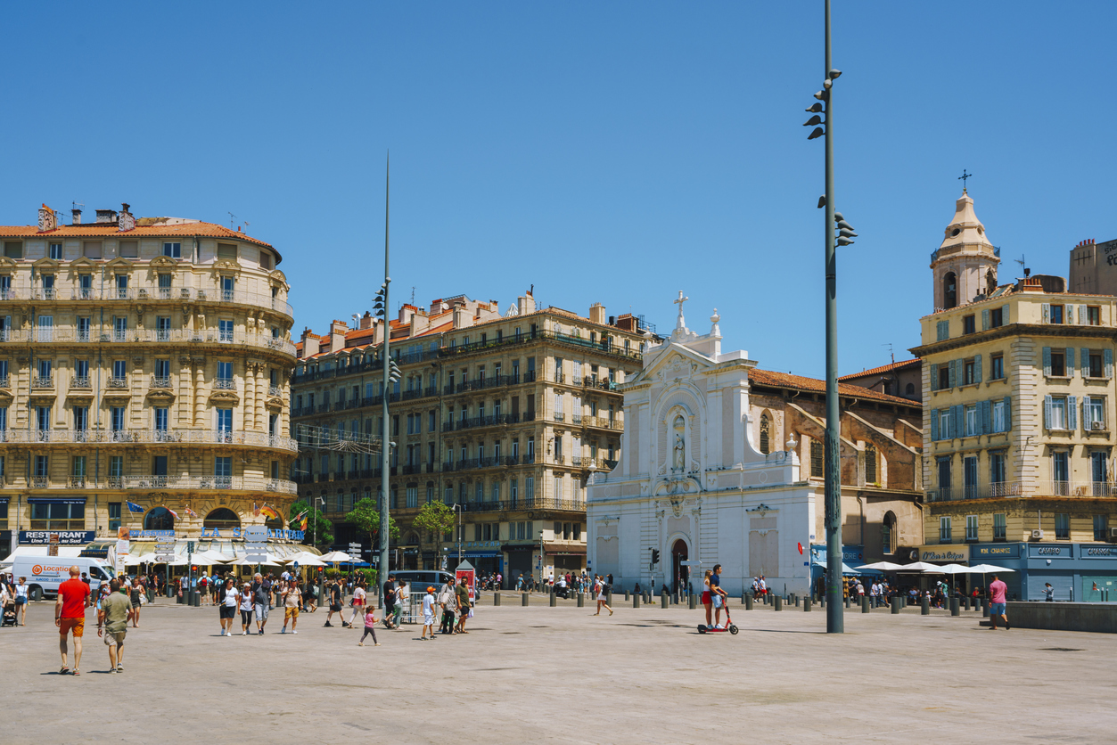 Belebter Platz am Vieux-Port in Marseille mit barocker weißer Stadtkirche, Haussmann-Fassaden, Spaziergängern und strahlend blauem Himmel.