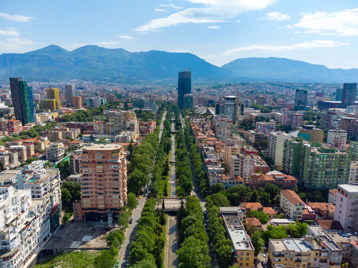 Luftaufnahme des Zentrums von Tirana mit einem breiten, baumbestandenen Grünzug entlang der Lana, modernem Hochhaus-Ensemble und Bergen des Dajti-Massivs im Hintergrund