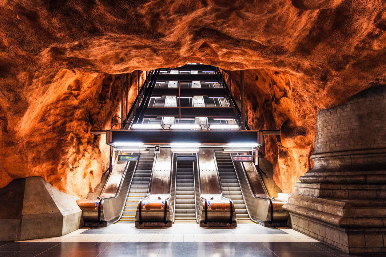 Stockholm U-Bahnhof Rådhuset, frontale Ansicht mit drei Rolltreppen unter rostroter, grob behauener Felsdecke, Beleuchtung über dem Aufgang, Bahnsteig im Vordergrund