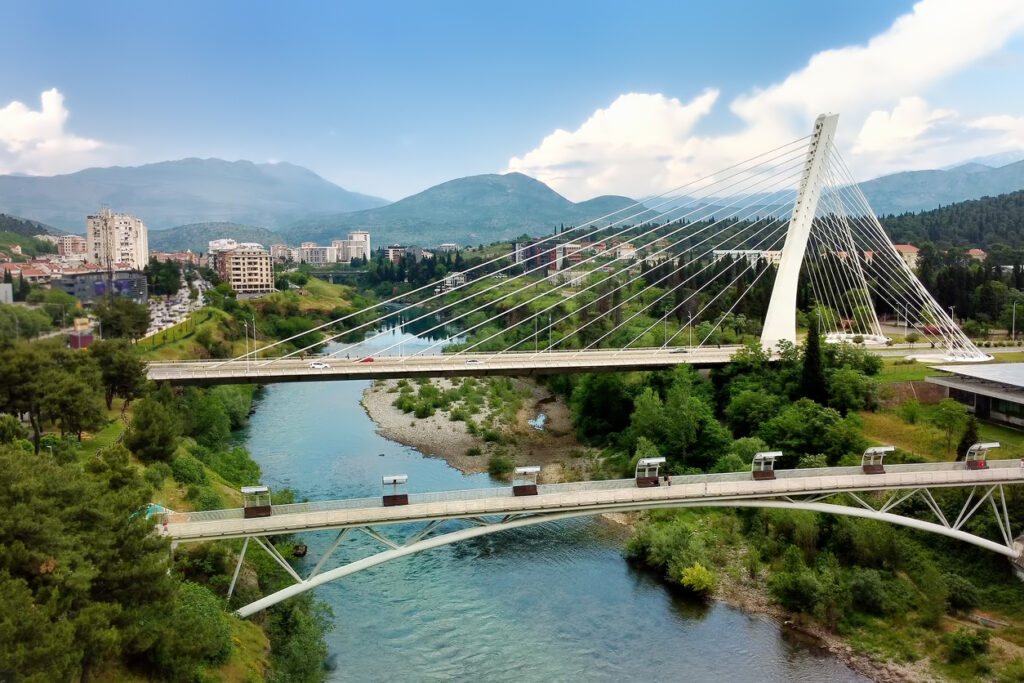 Blick auf die schrägseilverspannte Millennium-Brücke in Podgorica über dem türkisfarbenen Fluss Morača, darunter die geschwungene Moskva-Fußgängerbrücke, umgeben von viel Grün, Stadtvierteln und Bergen.