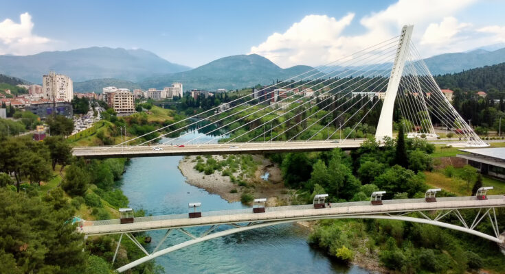 Blick auf die schrägseilverspannte Millennium-Brücke in Podgorica über dem türkisfarbenen Fluss Morača, darunter die geschwungene Moskva-Fußgängerbrücke, umgeben von viel Grün, Stadtvierteln und Bergen.