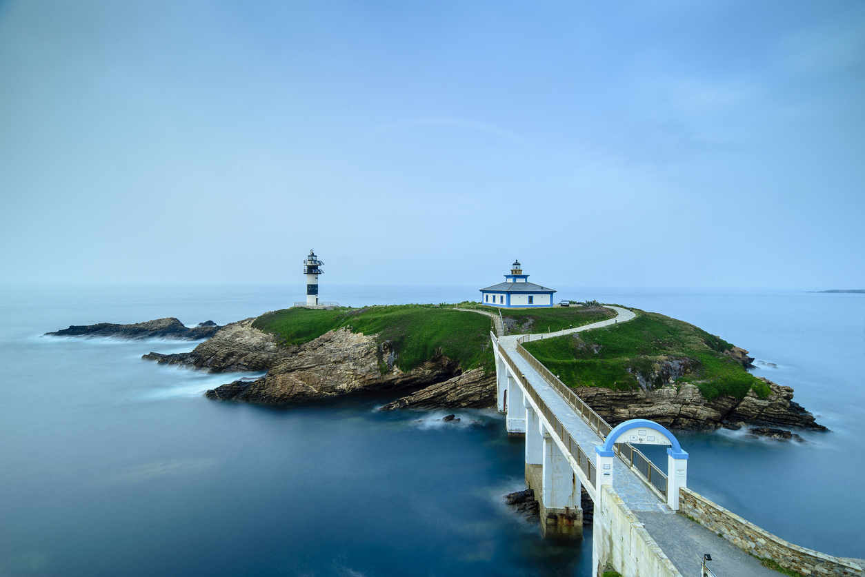 Malerischer Leuchtturm auf einer kleinen Insel in Ribadeo, verbunden durch eine Brücke mit dem Festland, umgeben vom blauen Meer.