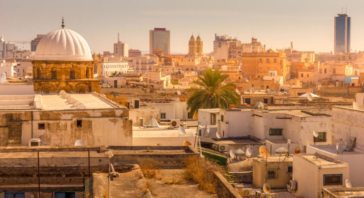 Blick über die Dächer der Medina von Tunis mit weißer Kuppelmoschee, Palmen und moderner Skyline im warmen Abendlicht.
