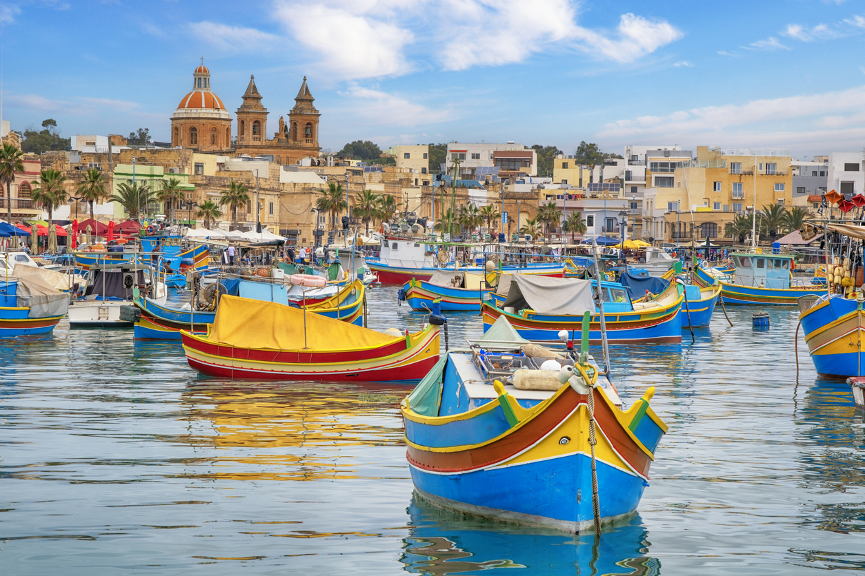 Bunte Fischerboote im Hafen von Marsaxlokk nahe Valletta, Malta, mit historischen Gebäuden im Hintergrund.