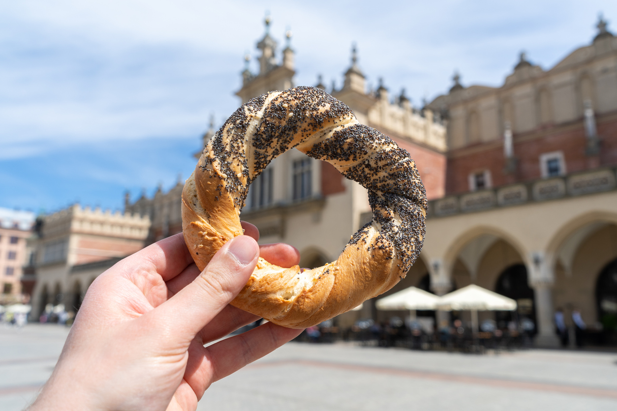 Hand hält einen Obwarzanek, das typische runde Gebäck aus Krakau, mit der Tuchhalle im Hintergrund.