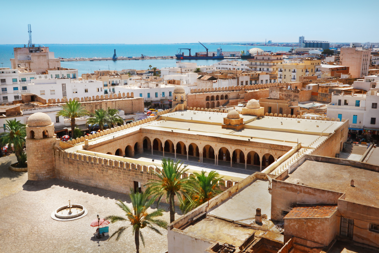 Luftaufnahme der Großen Moschee von Sousse mit Arkadenhof, Palmen, Stadtmauer und Blick auf den Hafen am Mittelmeer.