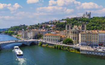 Panoramablick über die Saône in Lyon; ein Ausflugsschiff fährt unter dem Pont Bonaparte, rechts die Cathédrale Saint-Jean-Baptiste, darüber die Basilika Notre-Dame de Fourvière und der Fourvière-Turm auf dem Hügel.