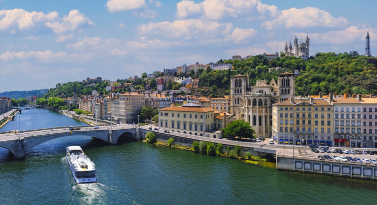 Panoramablick über die Saône in Lyon; ein Ausflugsschiff fährt unter dem Pont Bonaparte, rechts die Cathédrale Saint-Jean-Baptiste, darüber die Basilika Notre-Dame de Fourvière und der Fourvière-Turm auf dem Hügel.